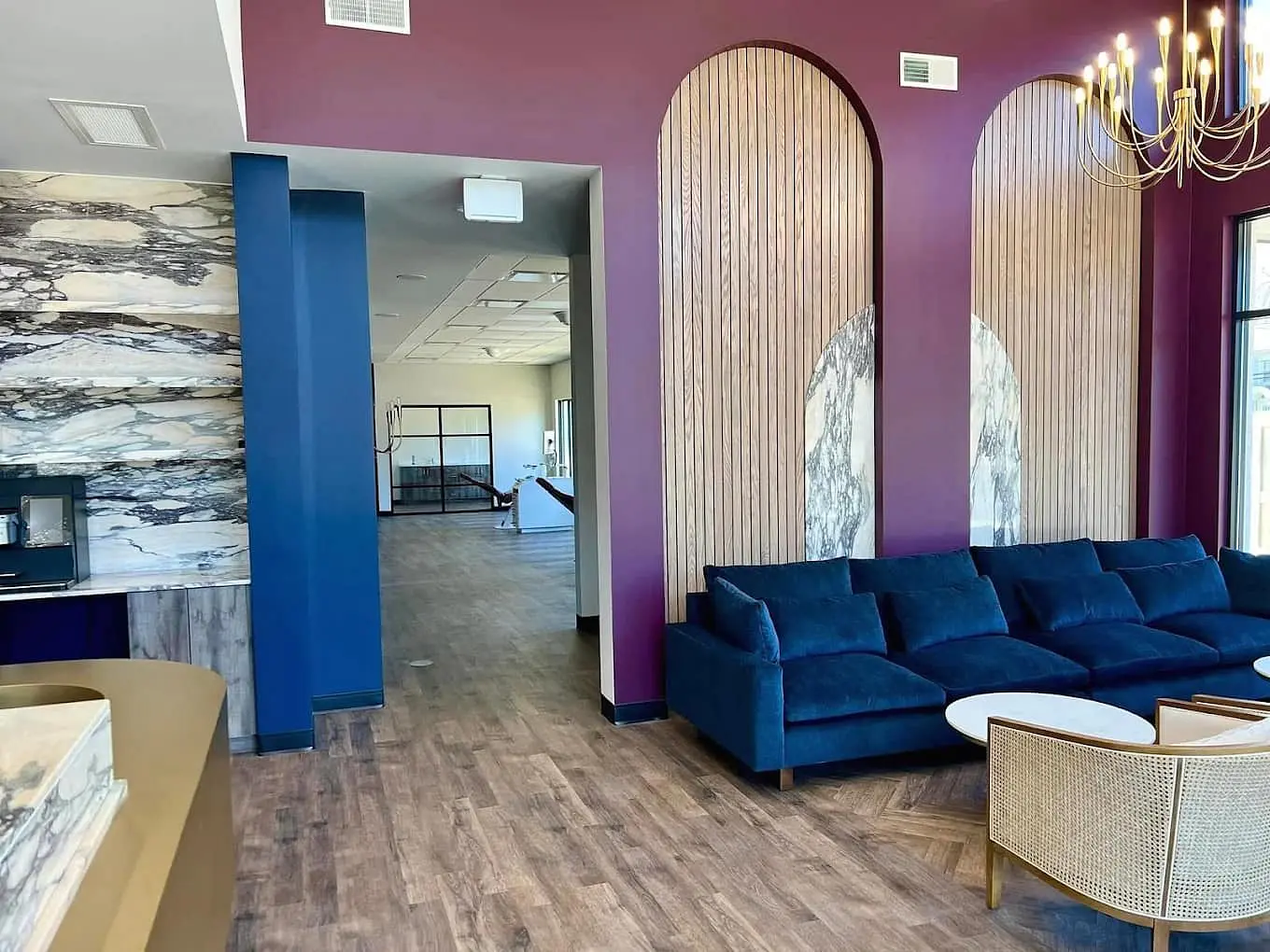 At Holt Orthodontics in Twinsburg, OH, the modern lounge area features a blue velvet sofa, wood accent walls, marble finishes, and a gold chandelier, seamlessly connecting to a spacious room with matching flooring and ceiling tiles.