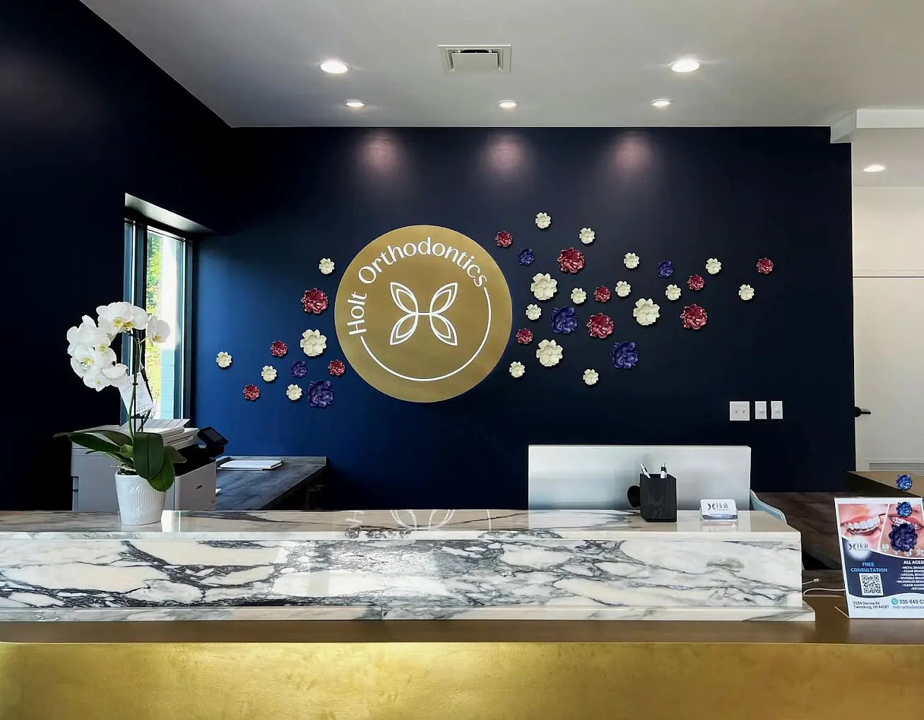 The reception area at Holt Orthodontics in Twinsburg, OH features a marble counter, flower decorations against a navy wall, and the clinic's gold logo displayed behind the desk.