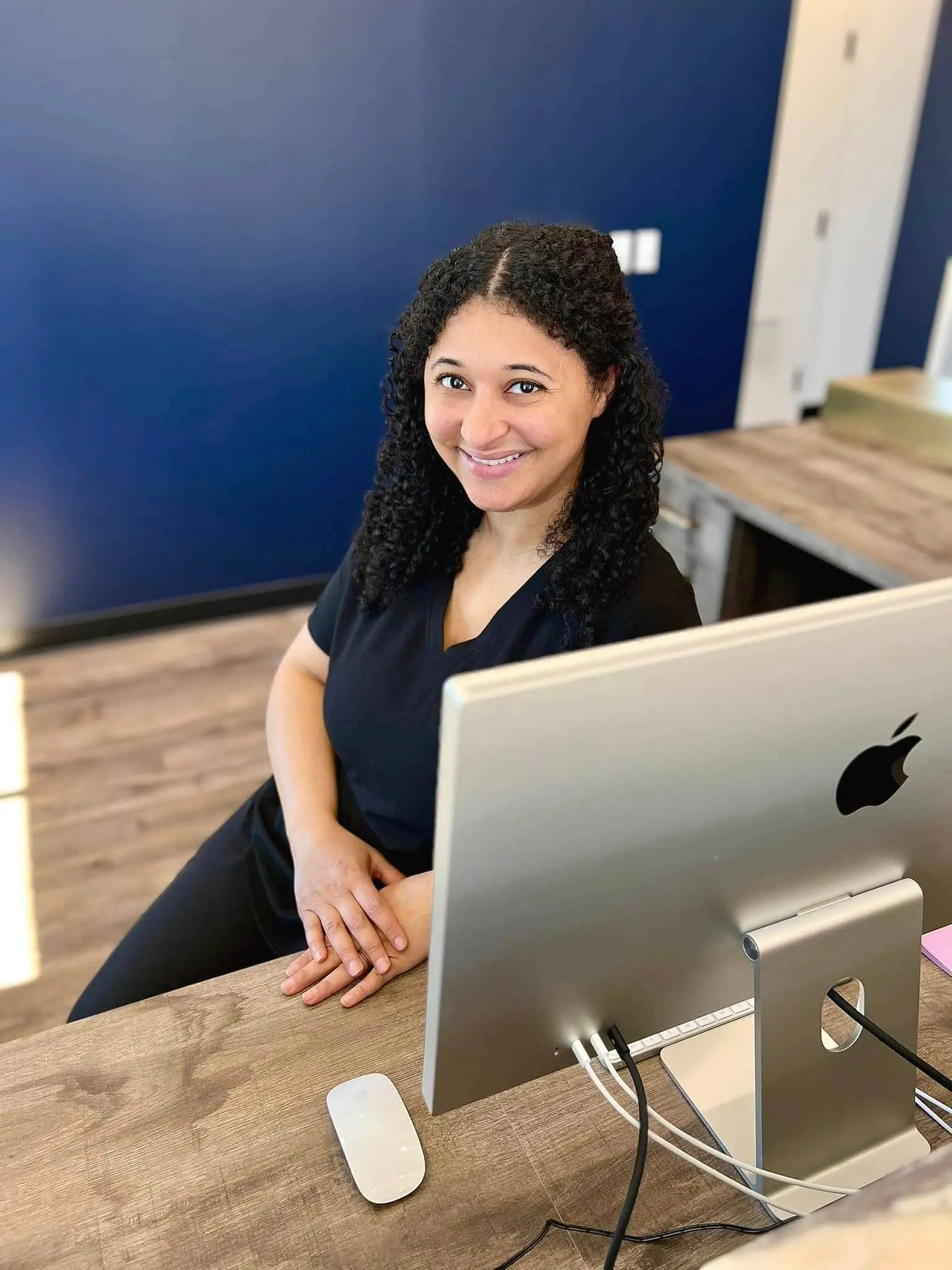 At Holt Orthodontics in Twinsburg, OH, a curly-haired orthodontics expert smiles at the camera while seated at a wooden desk with an Apple computer; the background includes a blue wall and a partially open door.