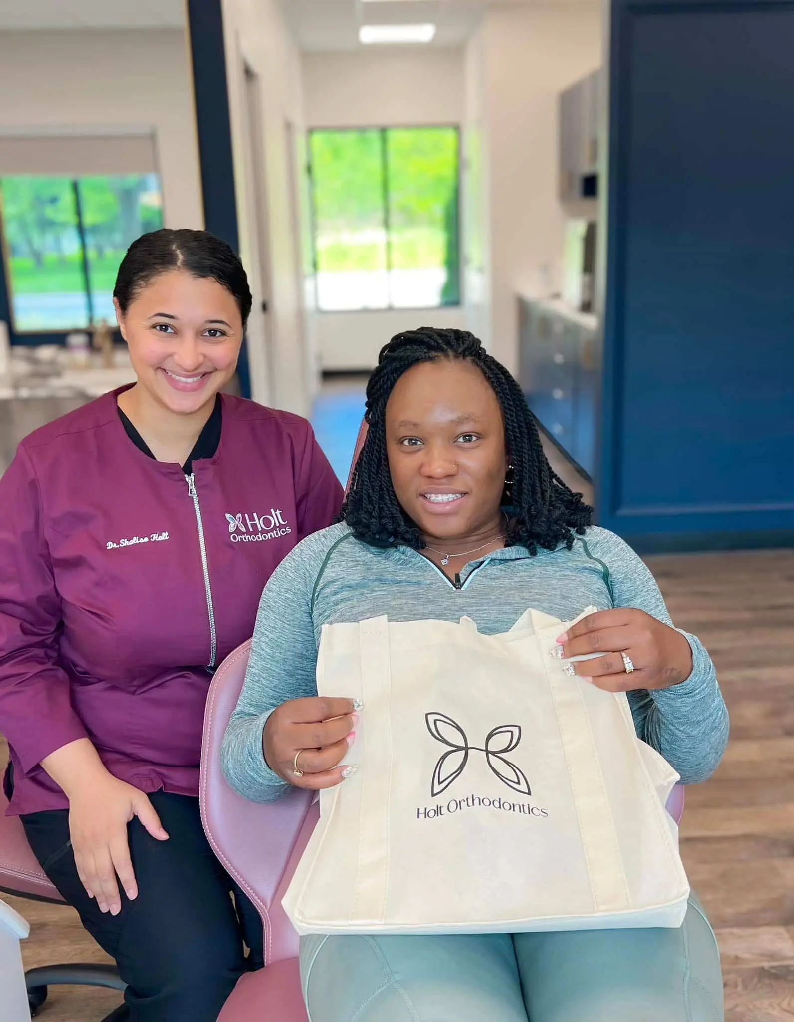 Inside a modern clinic in Twinsburg, OH, a woman sits in a dental chair holding a Holt Orthodontics tote bag beside Dr. Shalise Holt in a maroon uniform.