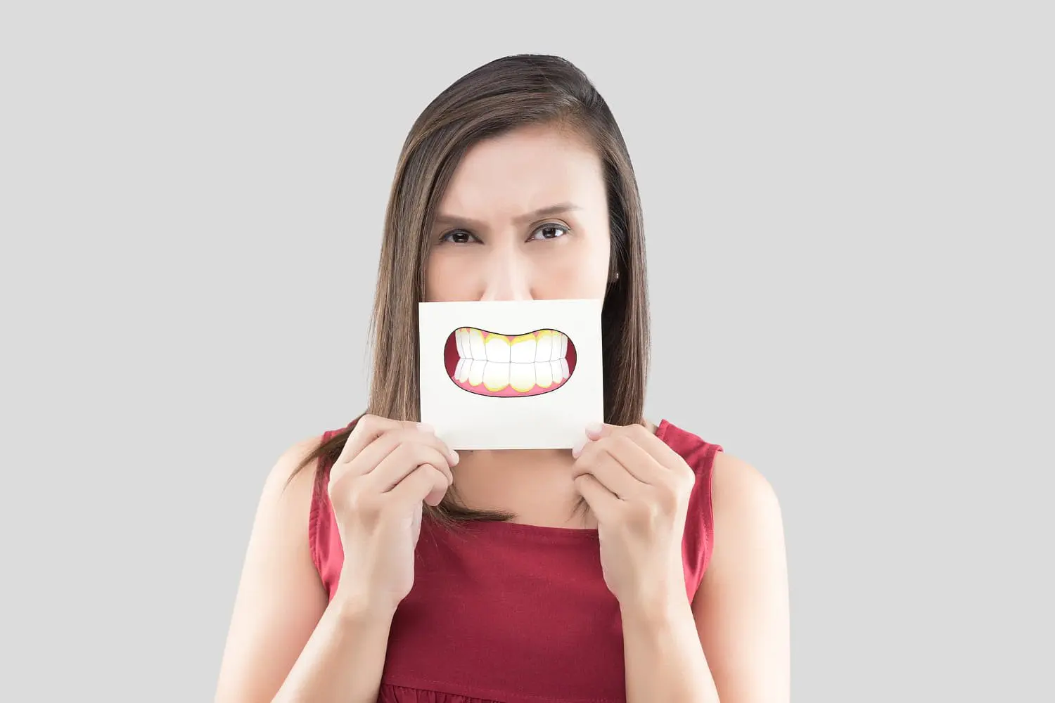 A woman in a red sleeveless top holds up a drawing of a large smiling mouth with an underbite in front of her face, set against a plain grey background—an image that captures the focus of Holt Orthodontics in Twinsburg, OH.