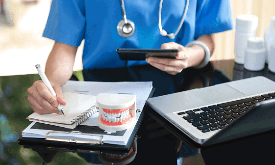 At Holt Orthodontics in Twinsburg, OH, a staff member in scrubs reviews orthodontic insurance plans for patients, taking notes beside a model of teeth, a laptop, and a tablet.