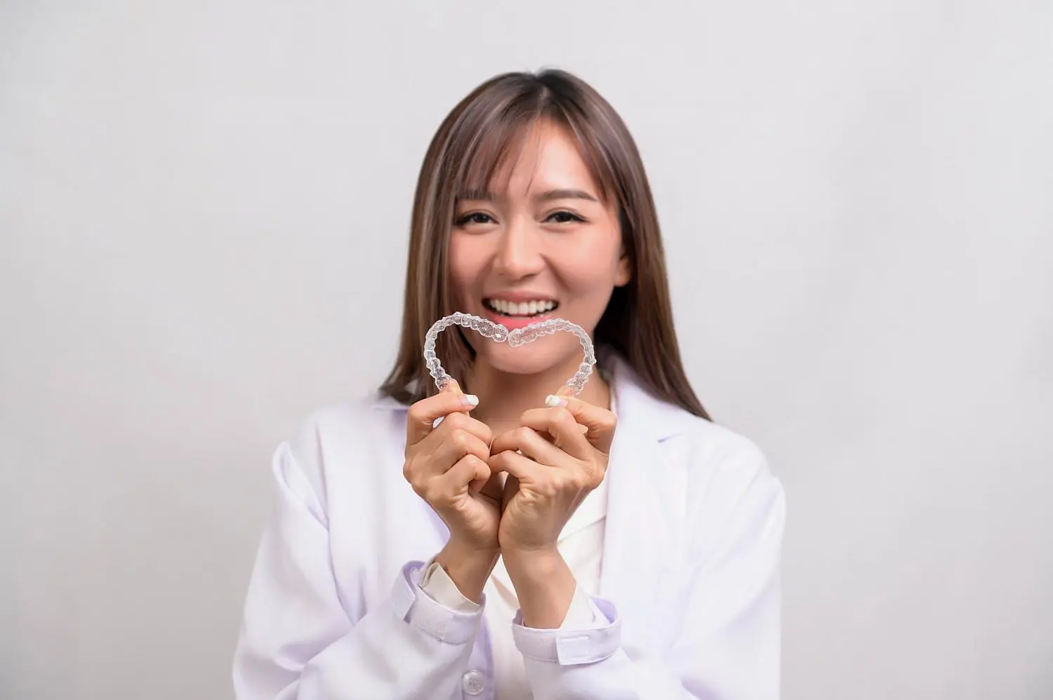 At Holt Orthodontics in Twinsburg, OH, a woman in a white coat smiles at the camera while holding two clear aligners shaped into a heart against a plain background.