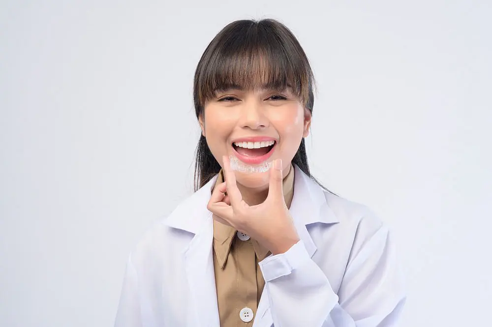 At Holt Orthodontics in Twinsburg, OH, a woman in a lab coat smiles and points to her teeth, displaying a clear dental aligner and showcasing the benefits and Invisalign cost for achieving a confident smile.
