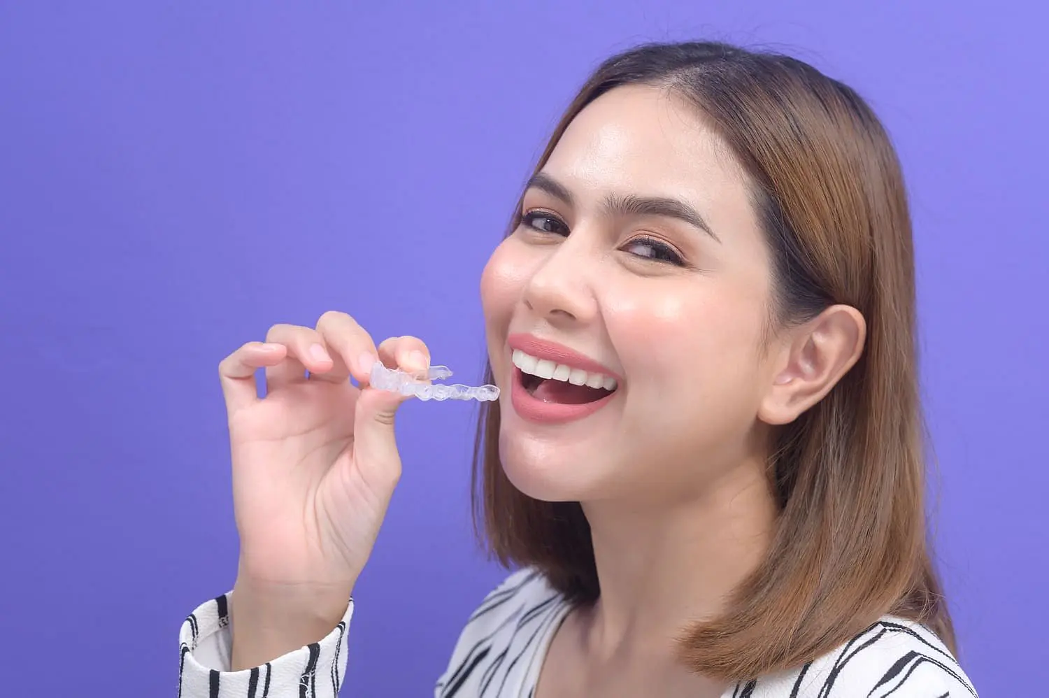 A woman smiles while holding Invisalign® Clear Aligners near her mouth, with a purple background, at Holt Orthodontics in Twinsburg, OH.
