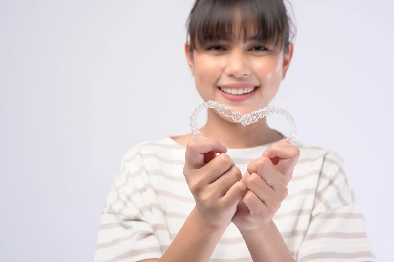 Smiling and holding an Invisalign clear dental aligner at chest level, a person in a striped shirt represents Holt Orthodontics in Twinsburg, OH, set against a plain background.