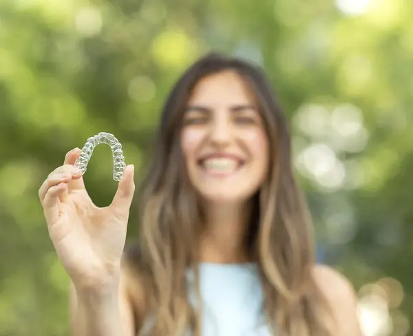 A woman smiles while holding a clear dental aligner in her outstretched hand, with a blurred green outdoor background, emphasizing that Invisalign at Holt Orthodontics in Twinsburg, OH may be covered by insurance.