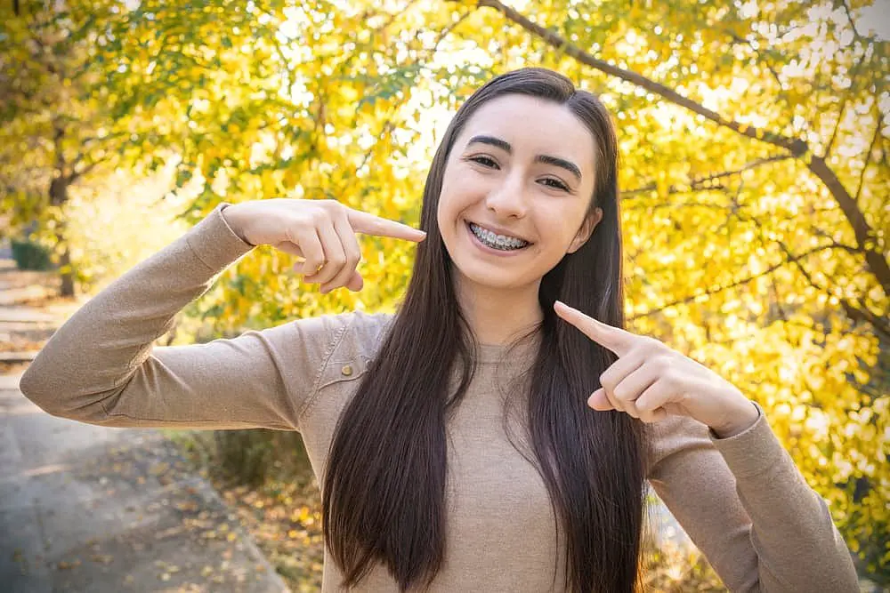 A young woman with long dark hair smiles outdoors, proudly pointing to her metal braces, with trees displaying yellow leaves behind her. This scene reflects the positive experiences patients have at Holt Orthodontics in Twinsburg, OH.