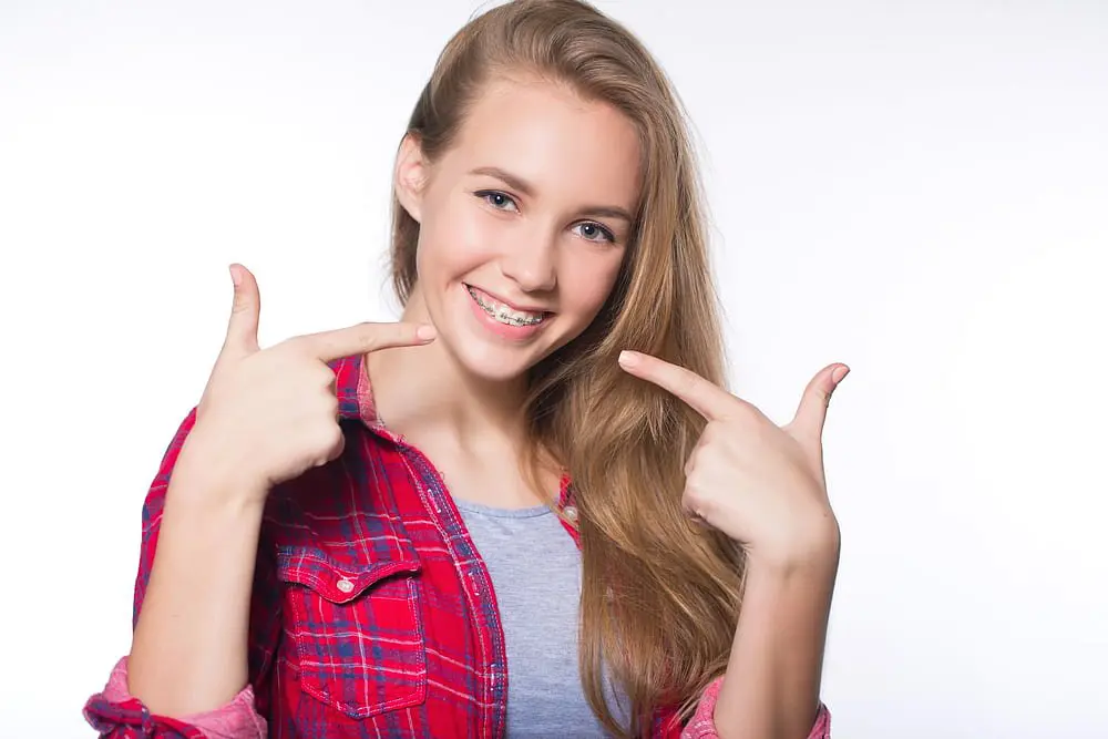 A teen girl in a red plaid shirt and gray top smiles proudly and points at her braces against a plain white background, showcasing the results from Holt Orthodontics in Twinsburg, OH.