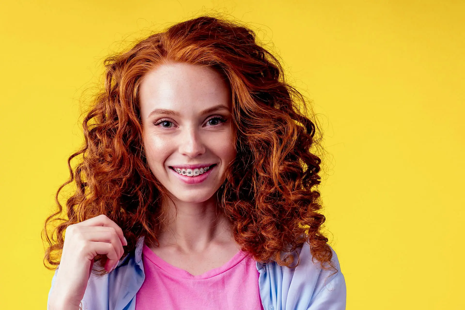 A young woman with curly red hair and metal braces smiles in front of a bright yellow background, sporting a pink shirt and light blue jacket—capturing the welcoming atmosphere at Holt Orthodontics in Twinsburg, OH.