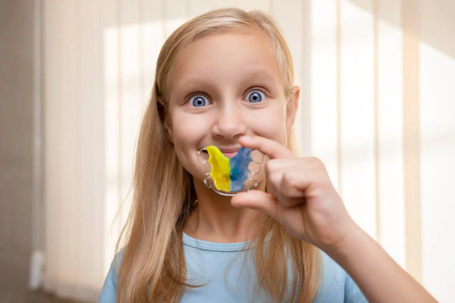 At Holt Orthodontics in Twinsburg, OH, a young girl with blonde hair smiles at the camera while holding up a colorful retainer mold, highlighting the fun experience of visiting a children’s orthodontist.