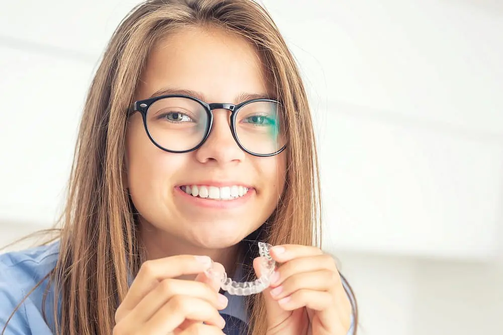 A smiling young person with glasses holds a clear aligner, showcasing the advantages of choosing Holt Orthodontics in Twinsburg, OH for children's clear aligner treatment.