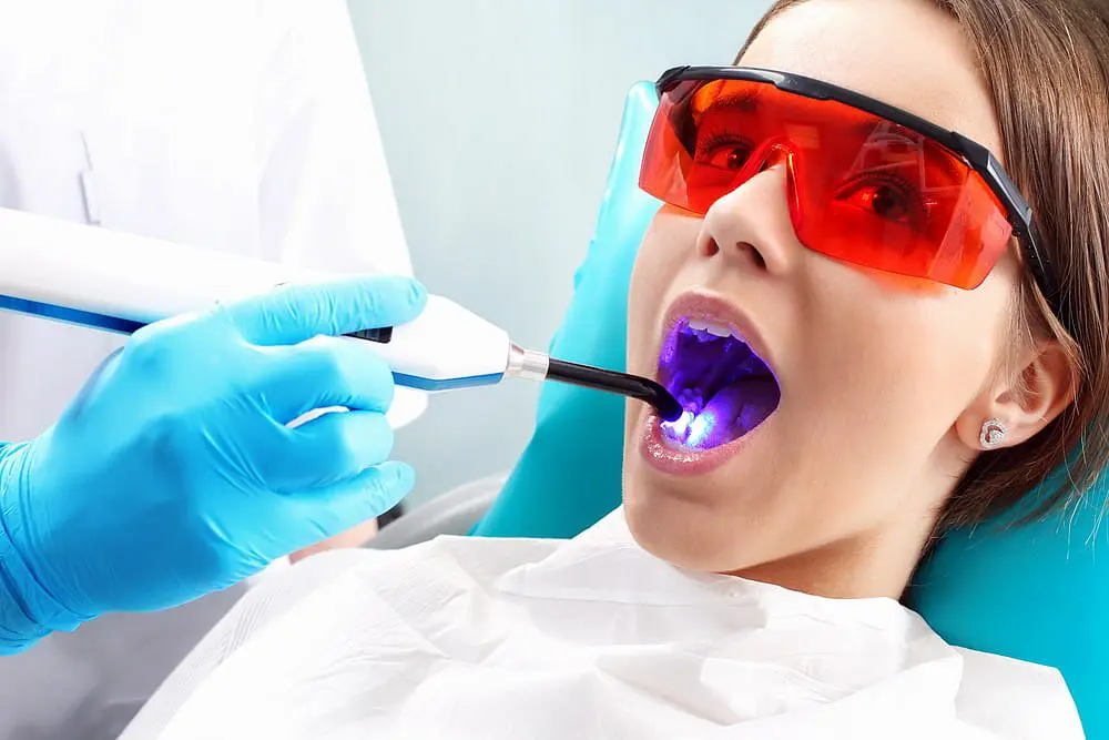 At Holt Orthodontics in Twinsburg, OH, a dental patient receives a teeth-whitening or curing treatment with a blue light device from a gloved dentist wearing red protective glasses, showcasing modern advances like Invisalign vs. braces.
