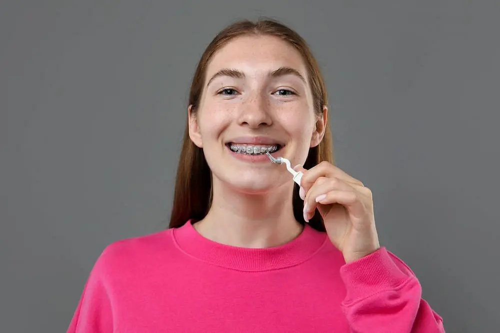 A young woman in a bright pink sweatshirt smiles while using a dental floss pick, illustrating daily care routines during the Invisalign vs Braces journey at Holt Orthodontics in Twinsburg, OH.