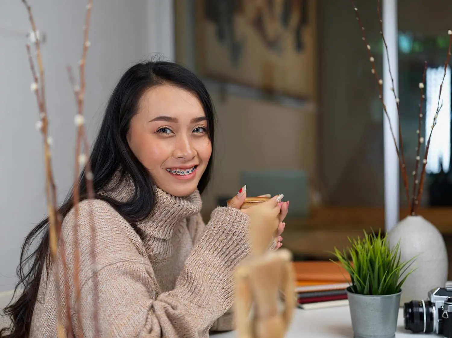A woman with long dark hair and braces smiles while enjoying a mug at a table, surrounded by plants and notebooks—an example of the benefits offered by Holt Orthodontics in Twinsburg, OH for adult orthodontic care.