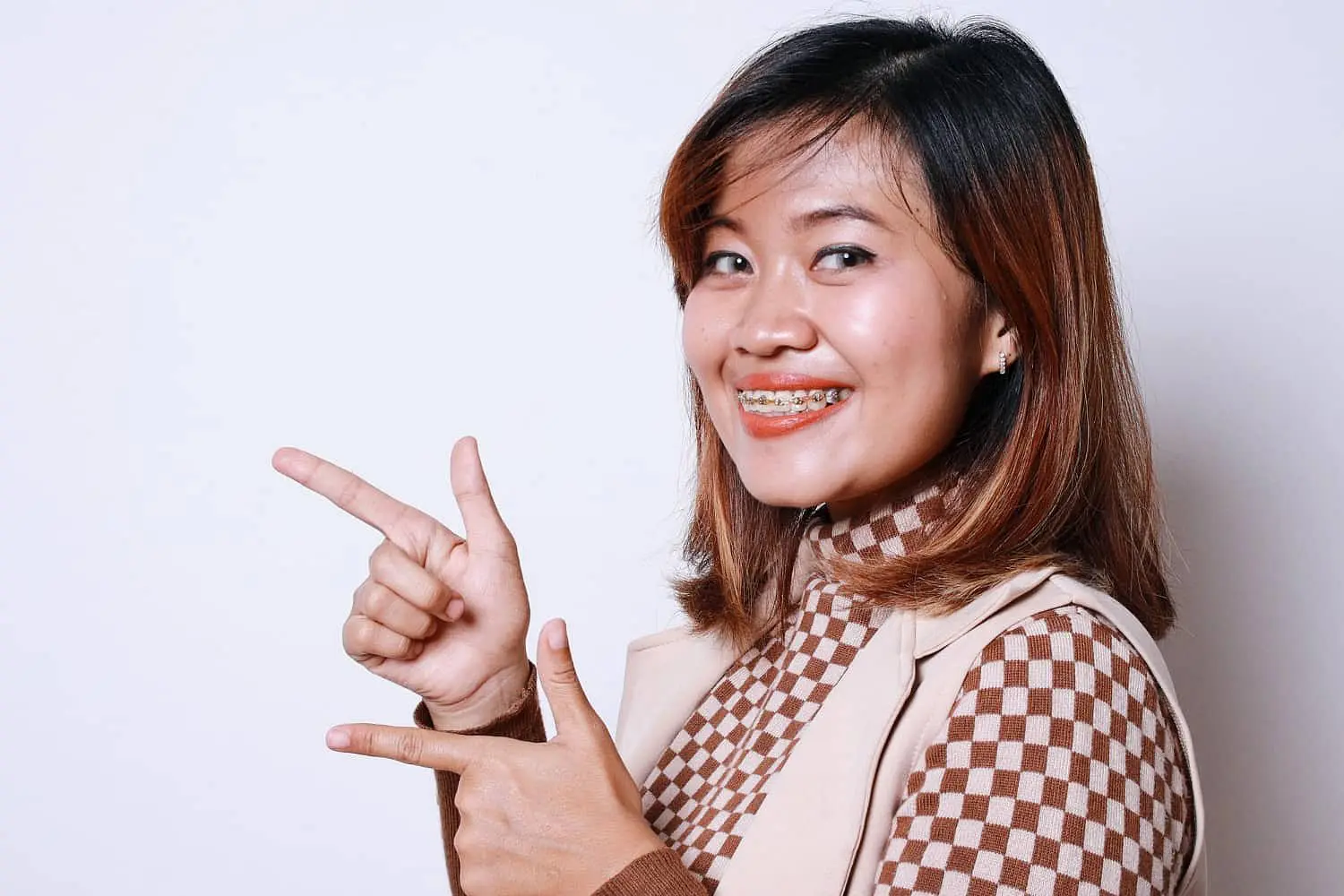 A woman with metal braces smiles and points to the side in front of a plain white background at Holt Orthodontics in Twinsburg, OH.