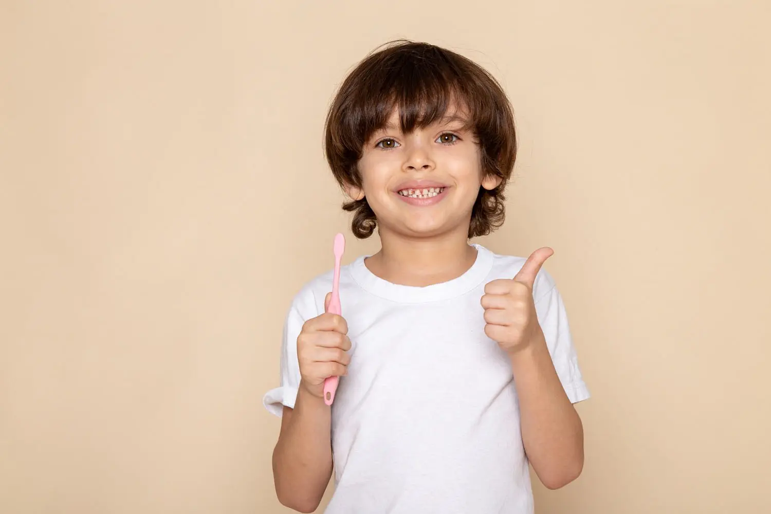 A smiling child in a white t-shirt holds a pink toothbrush and gives a thumbs up, ready for a checkup with the Children’s Orthodontist at Holt Orthodontics in Twinsburg, OH.