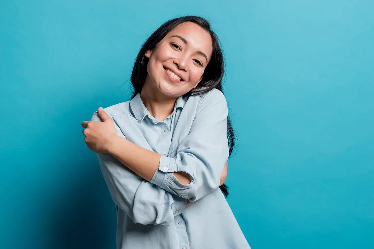 A woman in a light blue shirt with metal braces smiles and hugs herself against a solid blue background, representing Holt Orthodontics in Twinsburg, OH.