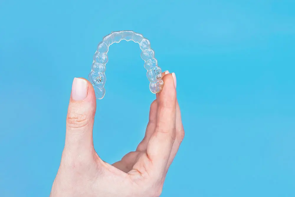 Against a blue background, a hand displays a clear dental aligner, highlighting the discreet design and value of Invisalign cost at Holt Orthodontics in Twinsburg, OH.