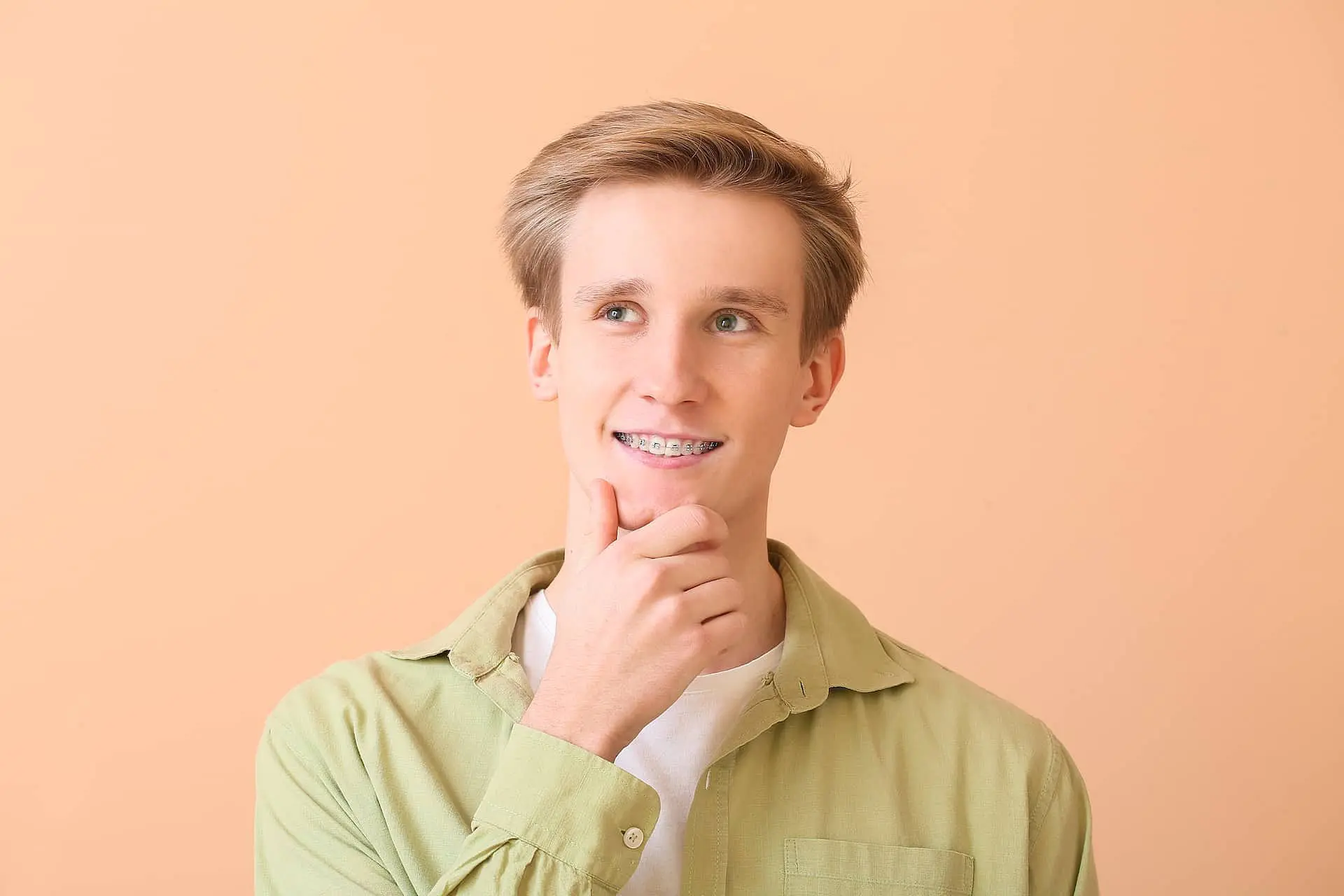 A young man with metal braces stands against a peach background, wearing a light green shirt over a white t-shirt and touching his chin while looking to the side. Holt Orthodontics in Twinsburg, OH can help you achieve your best smile.
