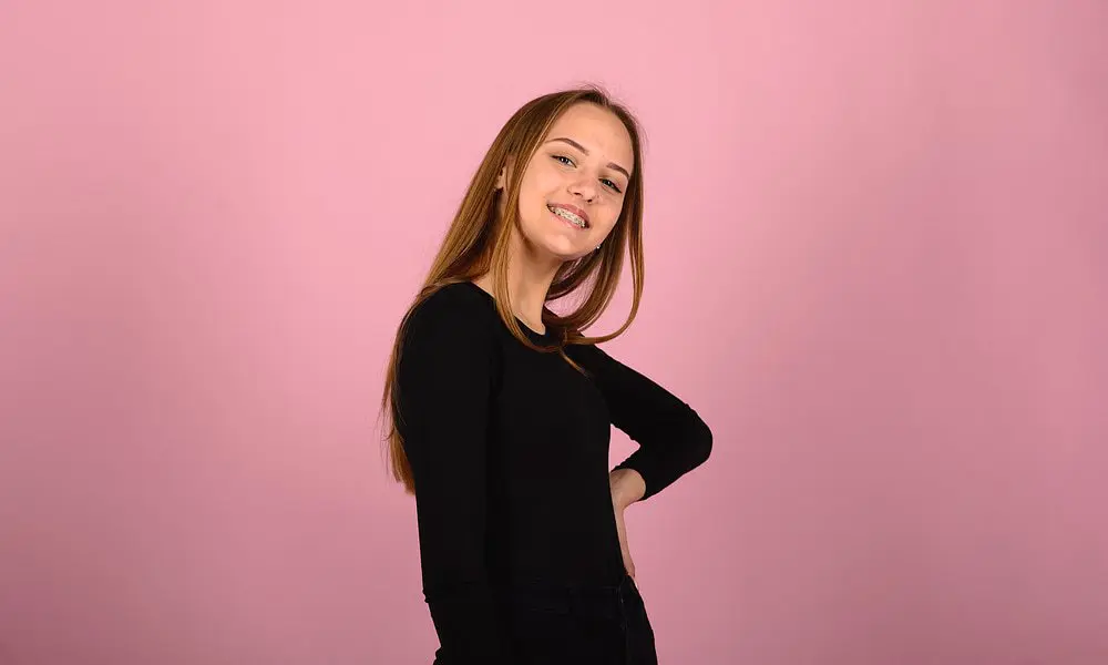 At Holt Orthodontics in Twinsburg, OH, a young woman with long straight hair and braces smiles against a plain pink background while wearing a black long-sleeve top.