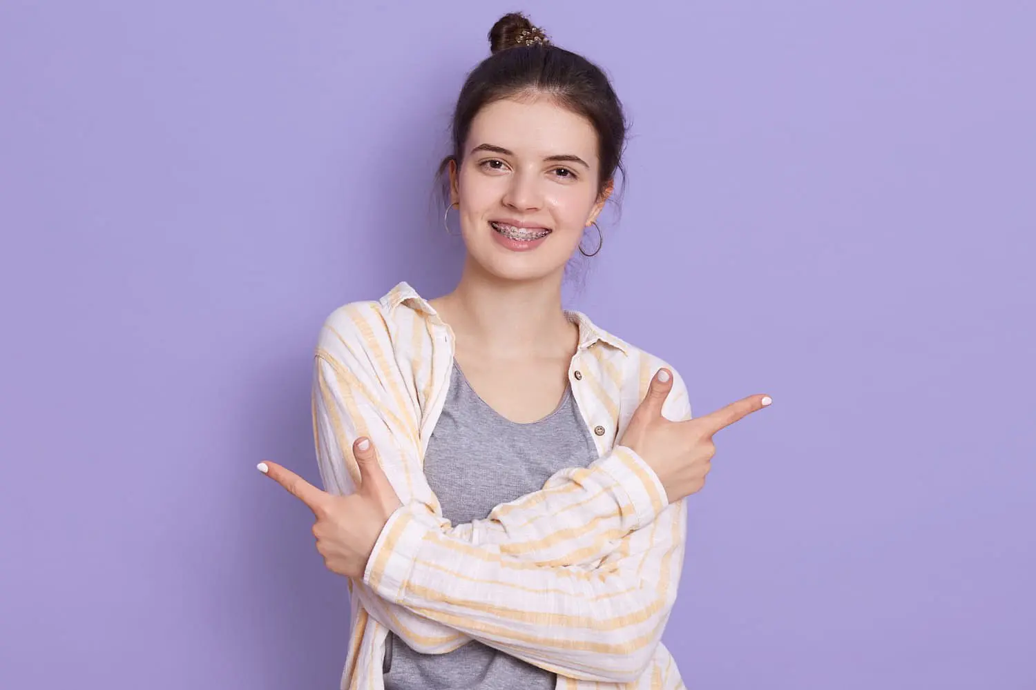 Holt Orthodontics A confident young woman with braces stands before a purple background, smiling with crossed arms and pointing both index fingers in opposite directions after discovering the affordable braces cost at Holt Orthodontics in Twinsburg, OH.