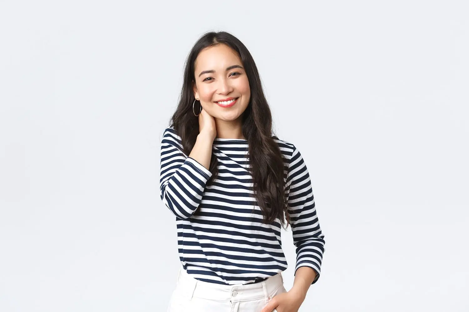 A woman in a black and white striped long-sleeve shirt stands against a plain background, smiling at the camera with her hand on her neck, subtly highlighting her charming overbite—an example of cases treated by Holt Orthodontics in Twinsburg, OH.