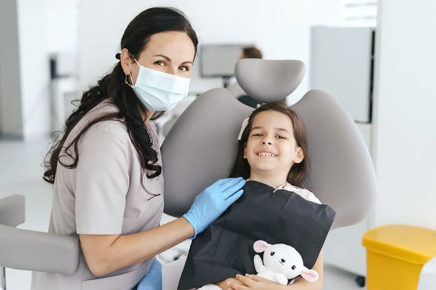 At Holt Orthodontics in Twinsburg, OH, a masked and gloved dentist sits beside a smiling young girl with her stuffed animal in a modern dental clinic, ready to discuss her new palatal expander.