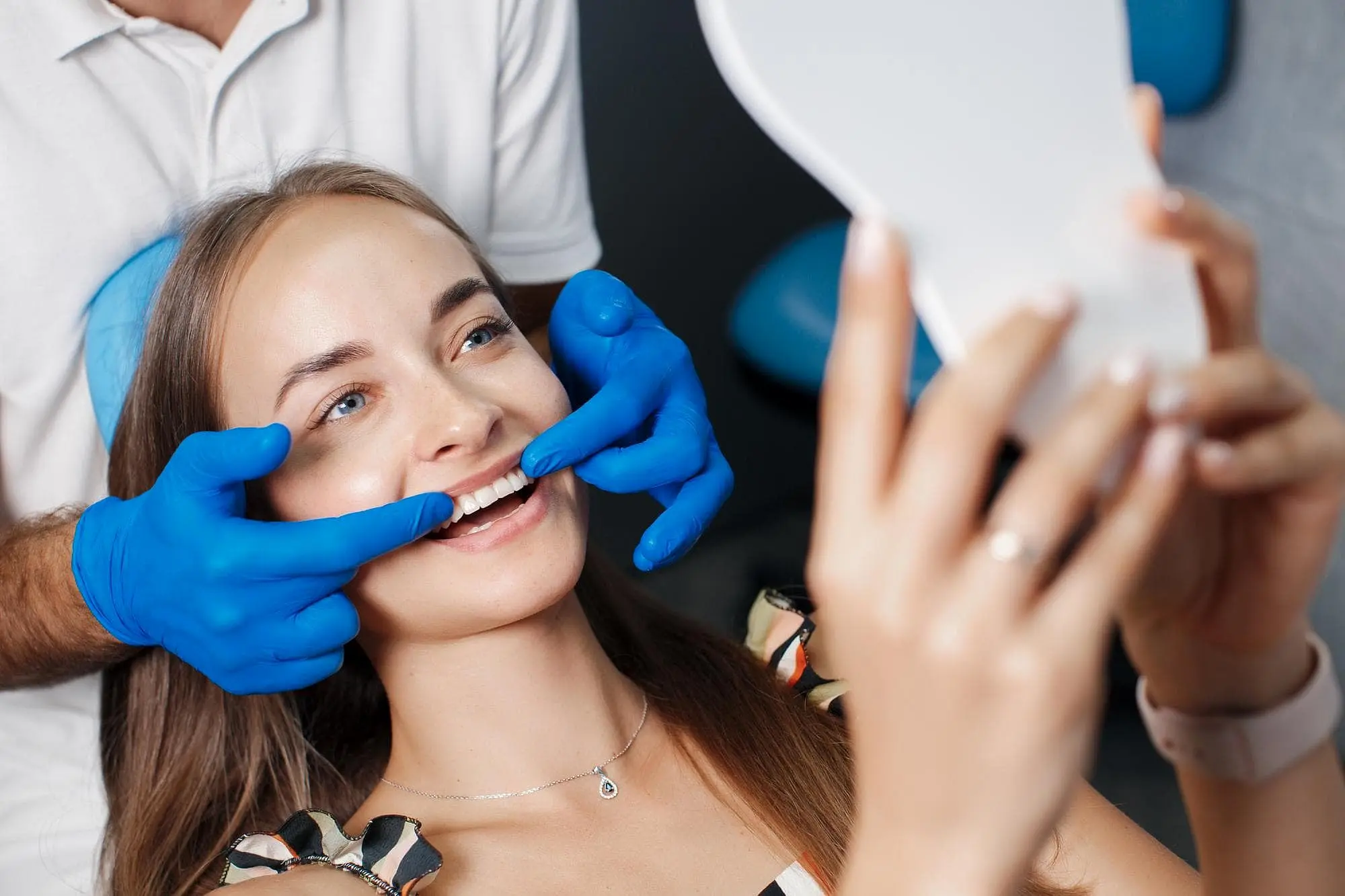 At Holt Orthodontics in Twinsburg, OH, a woman smiles at her reflection in a hand mirror as a dentist in blue gloves examines her teeth, showcasing the benefits of adult orthodontics.