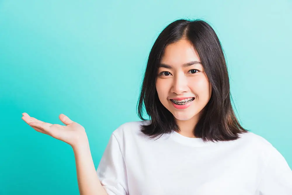 A young woman with straight dark hair, wearing metal braces and a white shirt, smiles and raises one hand against a plain light blue background at Holt Orthodontics in Twinsburg, OH.