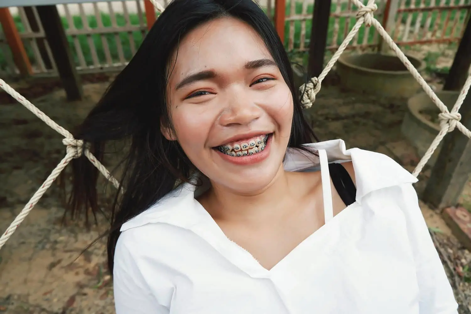 A young woman with long dark hair and braces, wearing a white shirt, smiles outdoors in front of a rope net, highlighting the benefits of adult orthodontics at Holt Orthodontics in Twinsburg, OH.
