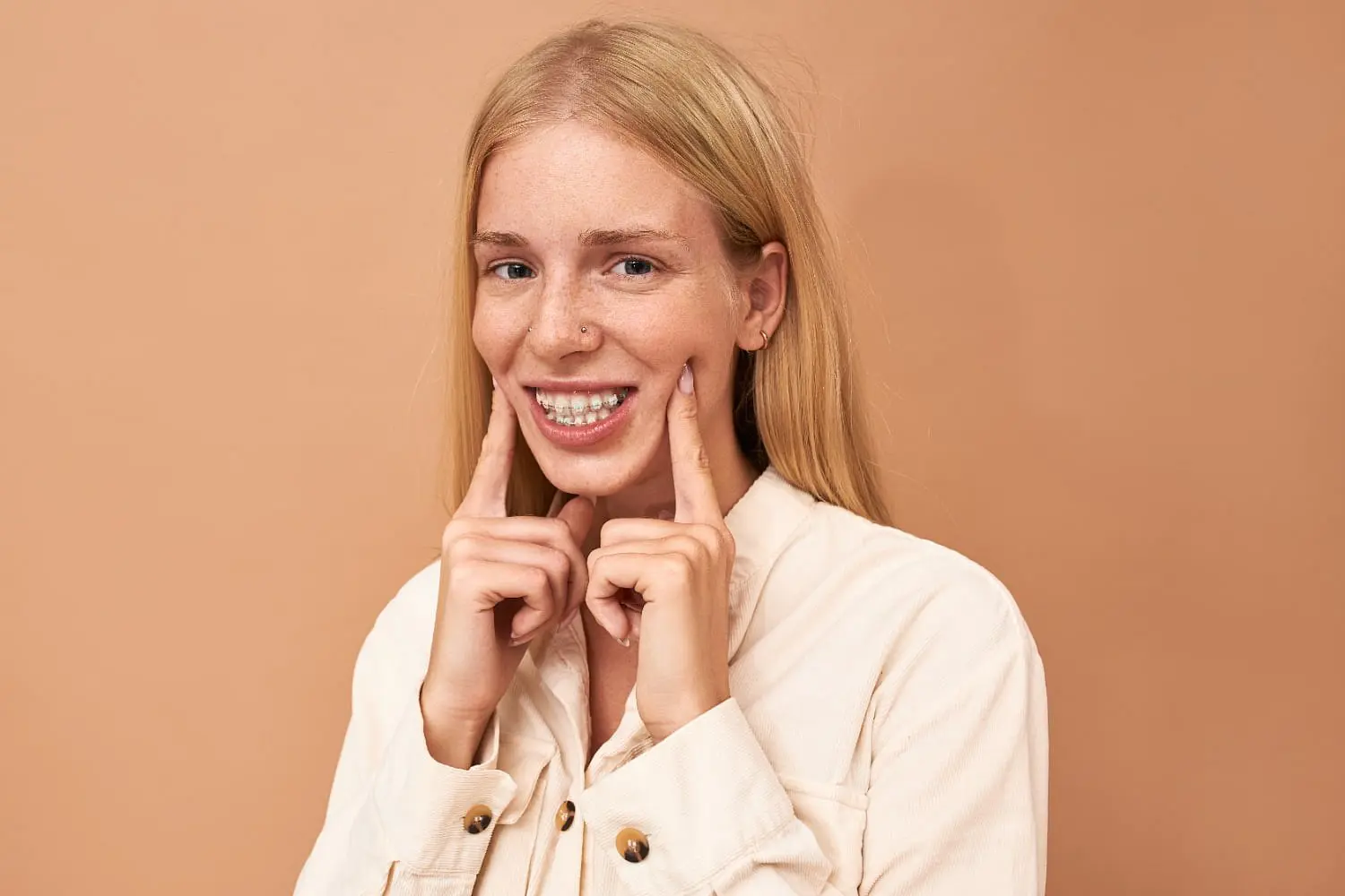 A person with long blonde hair and clear braces smiles and points to their teeth in front of a plain beige background at Holt Orthodontics in Twinsburg, OH.