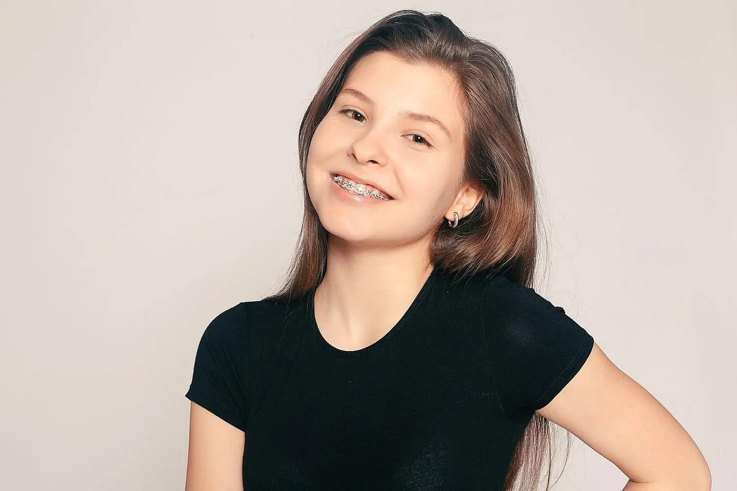 A teen girl with long brown hair, wearing a black t-shirt and braces, smiles confidently at the camera against a plain light background. This confidence reflects the healthy smiles achieved through Holt Orthodontics in Twinsburg, OH, which offers treatment options for both teens and adults.