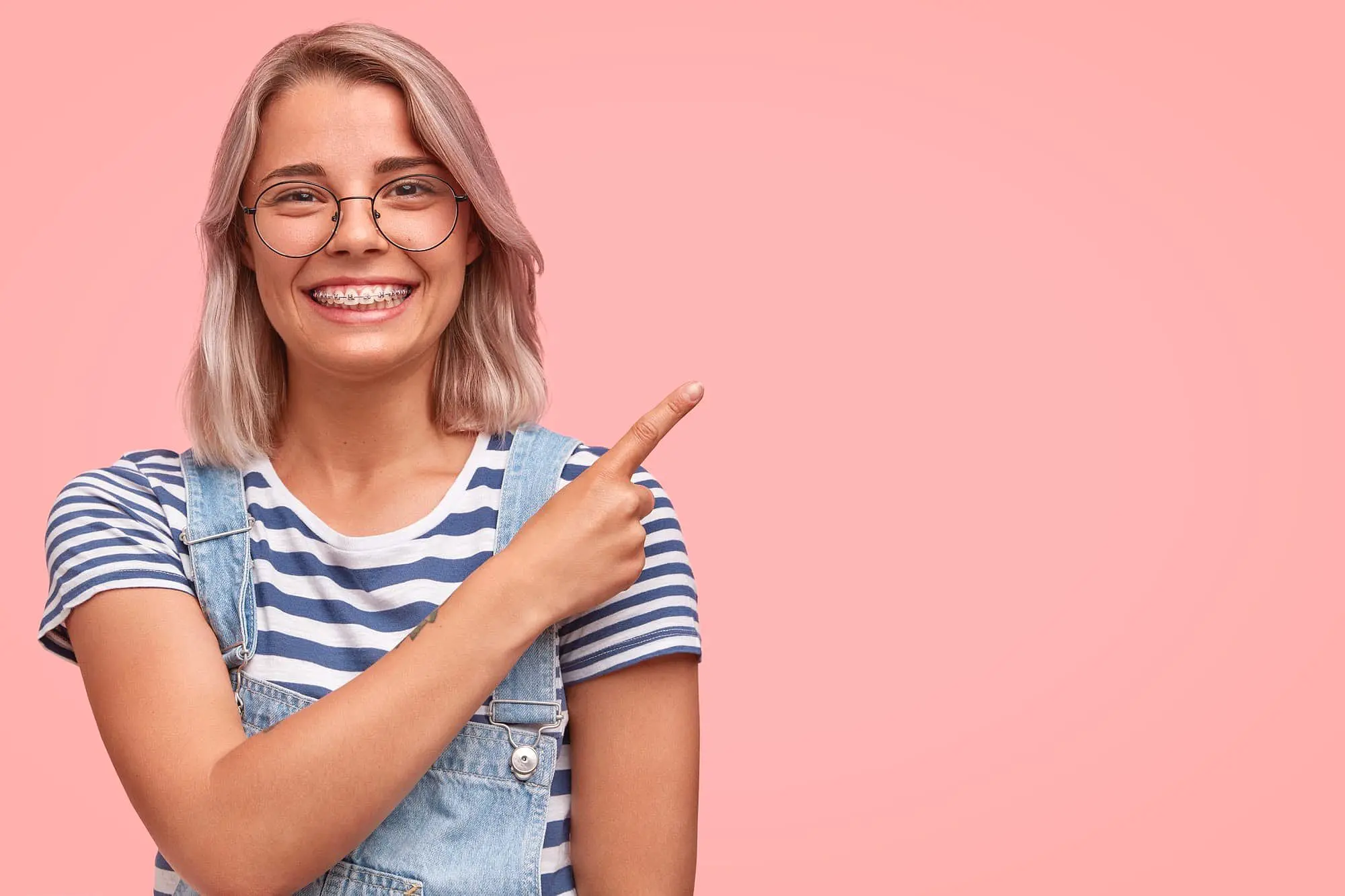 A smiling person with glasses and braces, dressed in a striped shirt and denim overalls, points right against a pink background—demonstrating the confidence that adult orthodontics from Holt Orthodontics in Twinsburg, OH can provide.