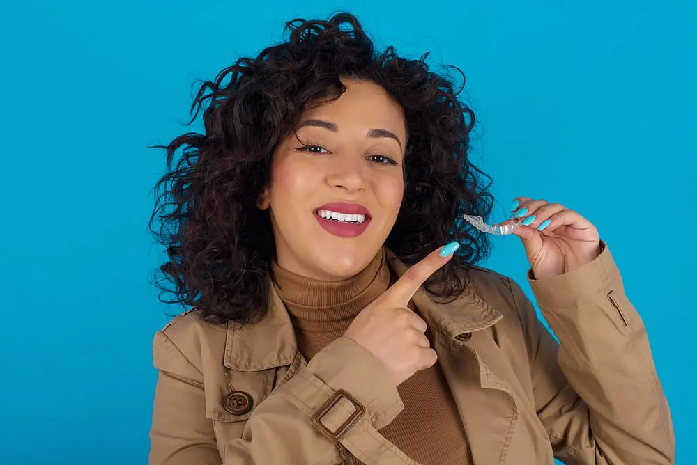 A woman with curly hair smiles and points to a clear aligner against a blue background, showcasing the benefits of an Invisalign Payment Plan offered by Holt Orthodontics in Twinsburg, OH.