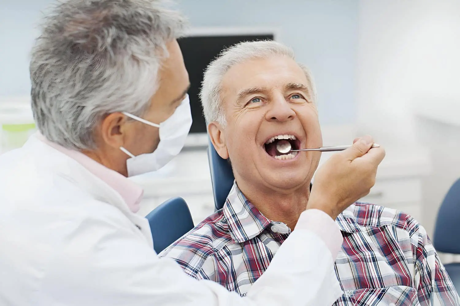 At Holt Orthodontics in Twinsburg, OH, a dentist wearing a mask uses a dental mirror to examine an older man’s teeth, assessing his oral health and discussing potential jaw surgery while the patient sits with his mouth open.