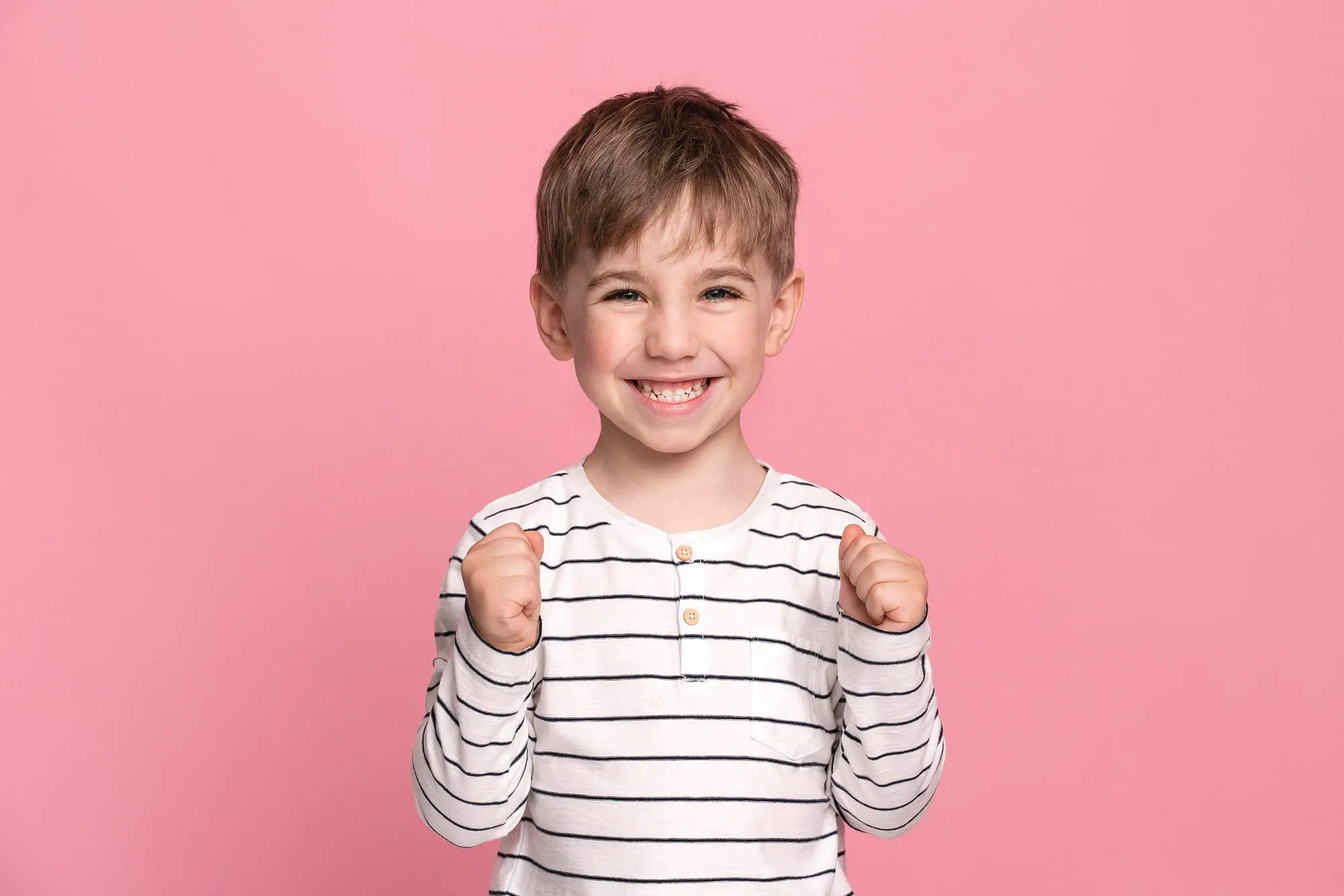 A smiling young boy in a white and black striped shirt stands against a pink background, proudly holding up his fists to show his confidence after getting affordable braces at Holt Orthodontics in Twinsburg, OH.