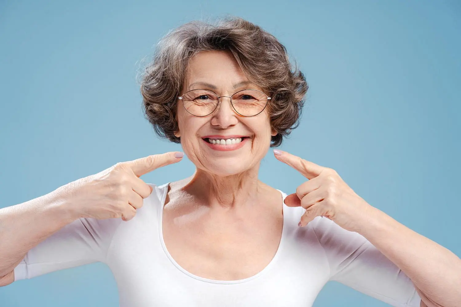 A smiling older woman with short hair and glasses points to her teeth against a blue background, highlighting the benefits of orthodontic insurance plans offered by Holt Orthodontics in Twinsburg, OH.