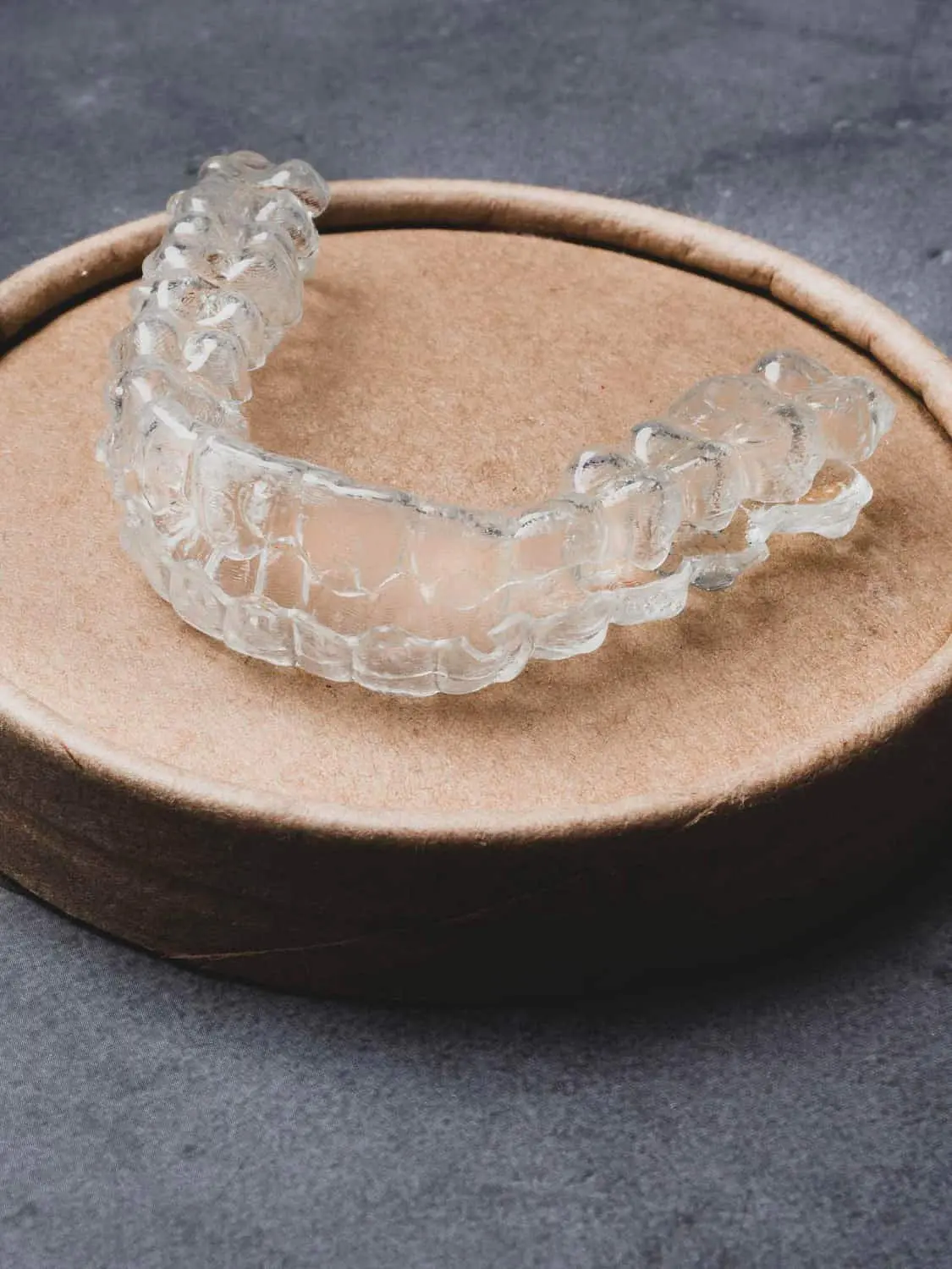 A transparent dental aligner from Holt Orthodontics in Twinsburg, OH, offered with an Invisalign Payment Plan, rests on a round brown container against a gray background.