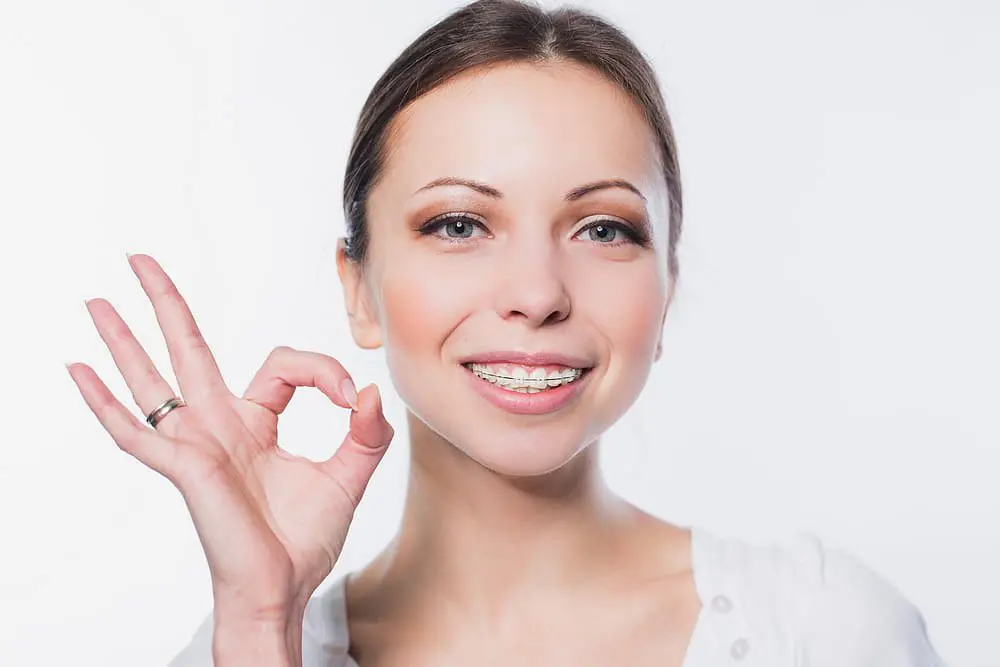 A woman with clear ceramic braces smiles at the camera and gives an "OK" gesture with her right hand, showcasing her treatment at Holt Orthodontics in Twinsburg, OH.