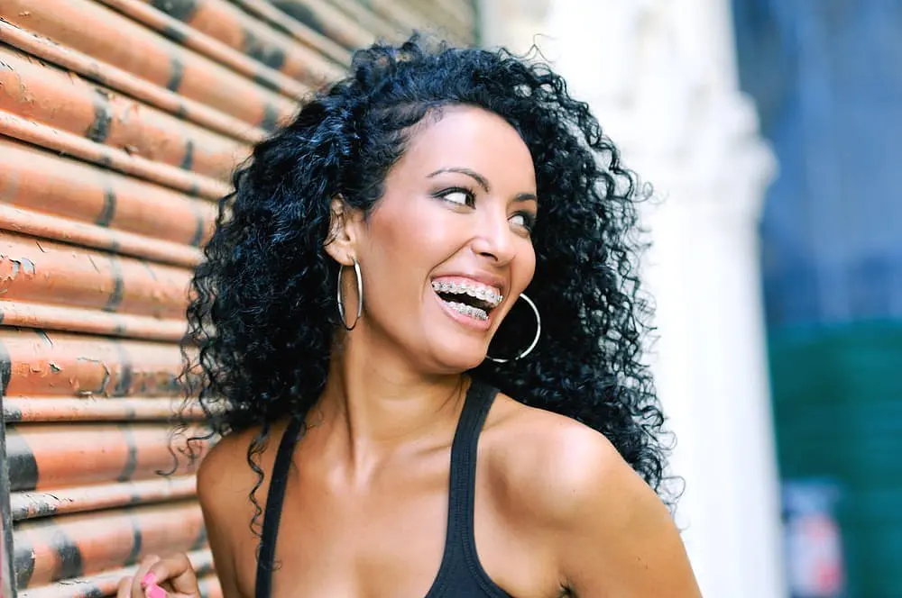 A woman with curly hair and adult braces smiles against a corrugated metal wall, wearing hoop earrings and a black top at Holt Orthodontics in Twinsburg, OH.