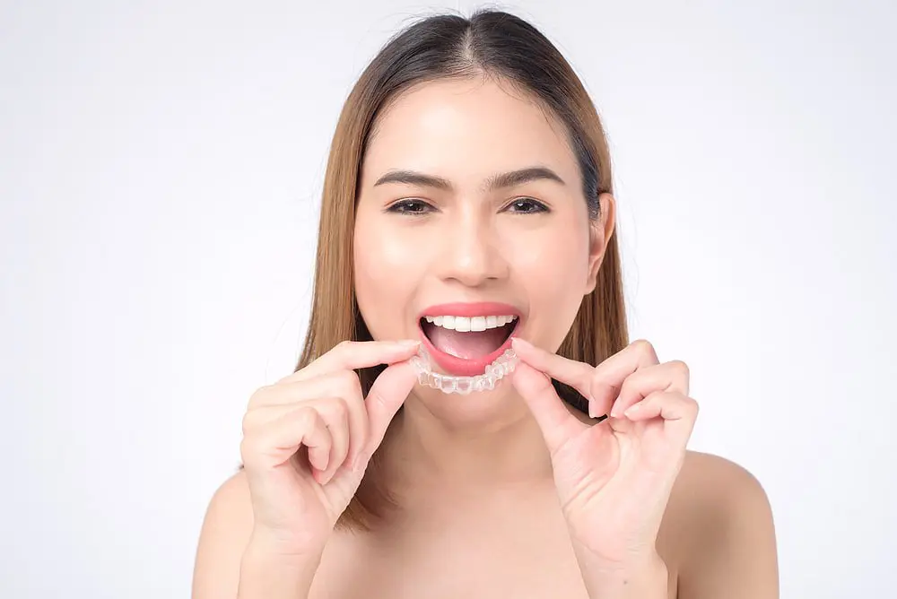 A woman with straight brown hair smiles while placing Invisalign® Clear Aligners on her upper teeth at Holt Orthodontics in Twinsburg, OH.