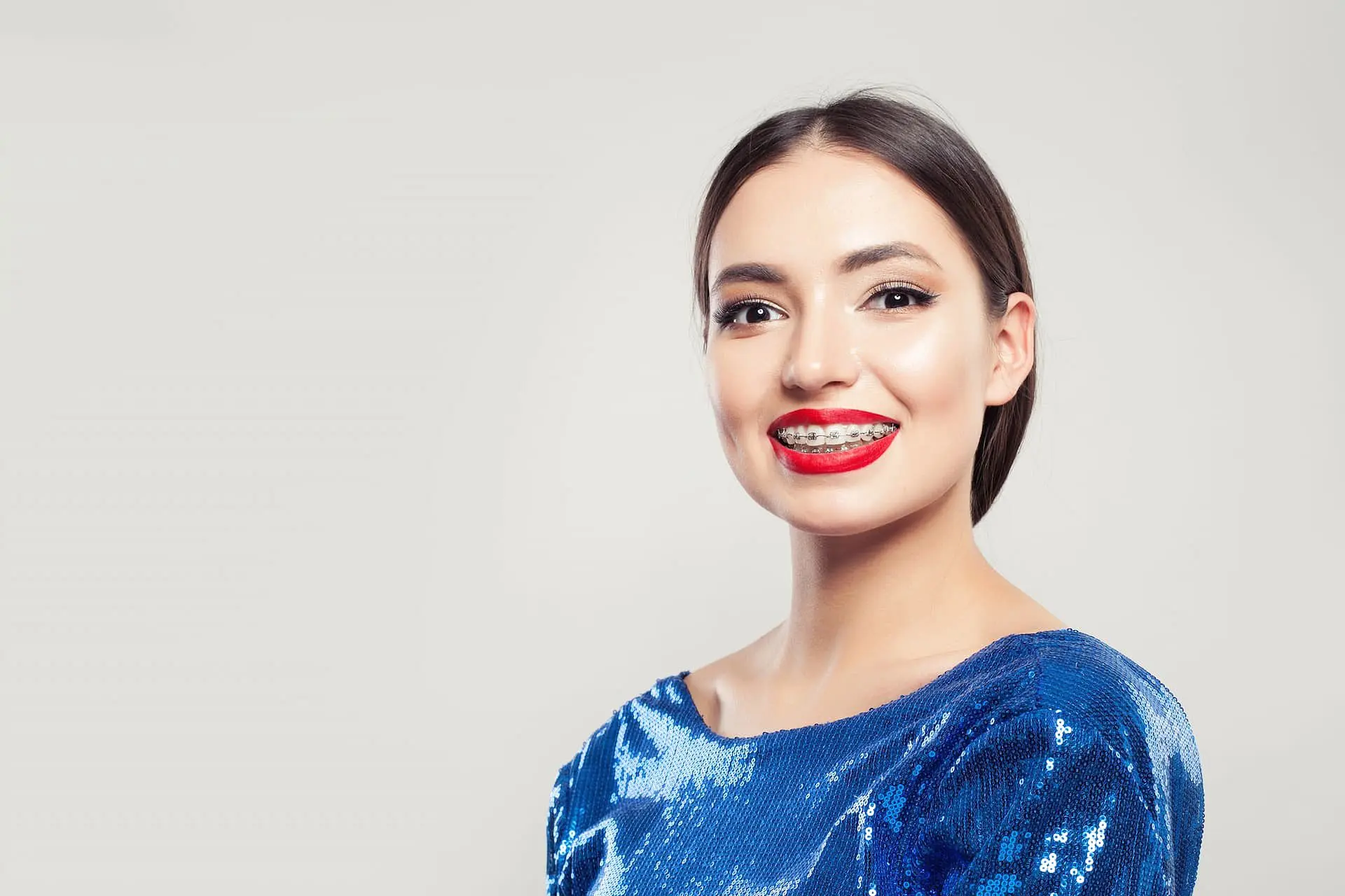 A woman with straight dark hair, wearing a blue sequin top and red lipstick, confidently smiles to reveal adult braces—proudly showcasing her results from Holt Orthodontics in Twinsburg, OH—against a light grey background.