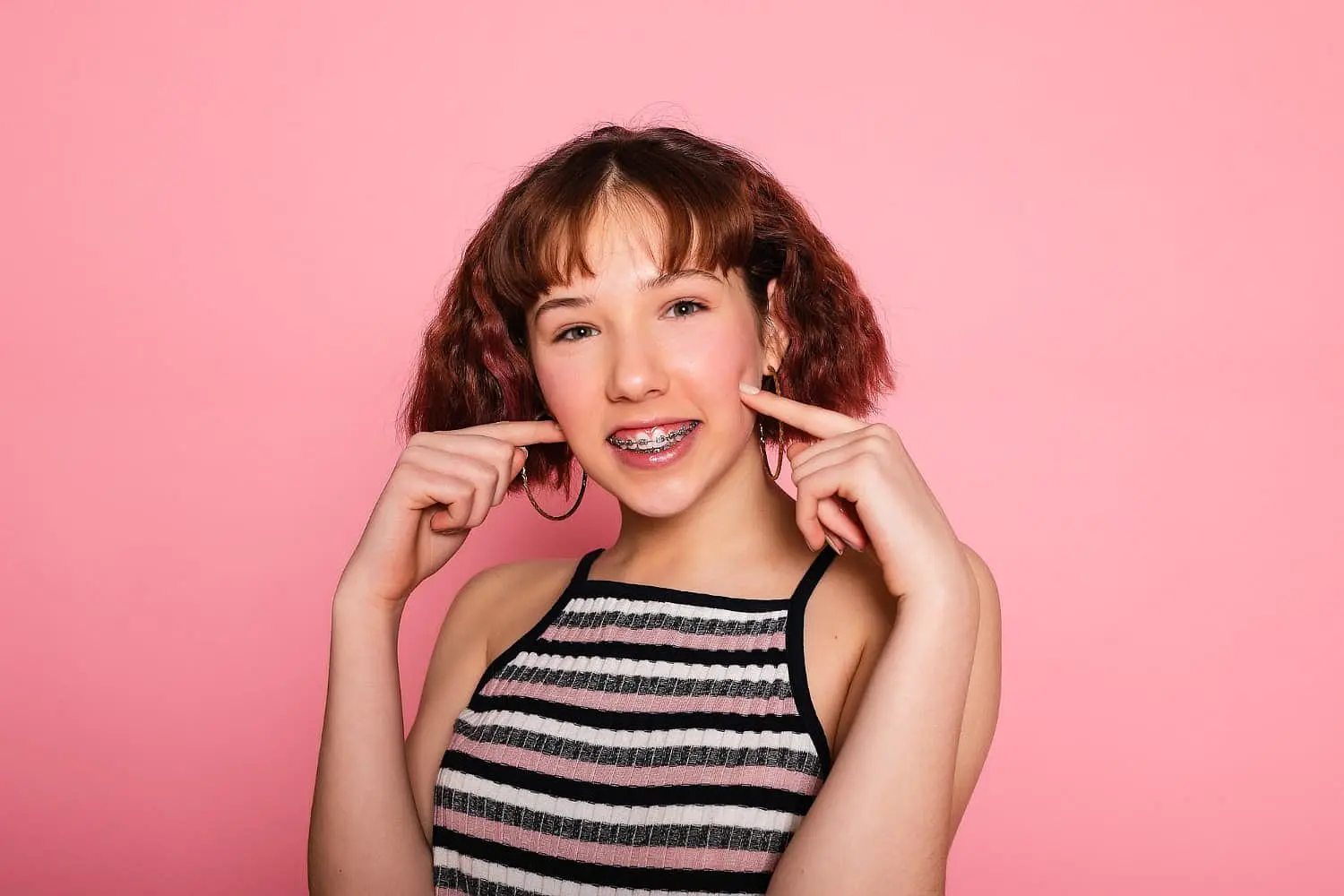 A smiling young person with braces and short brown hair points to their cheeks against a plain pink background, reflecting the happiness of choosing Invisalign vs Braces at Holt Orthodontics in Twinsburg, OH.