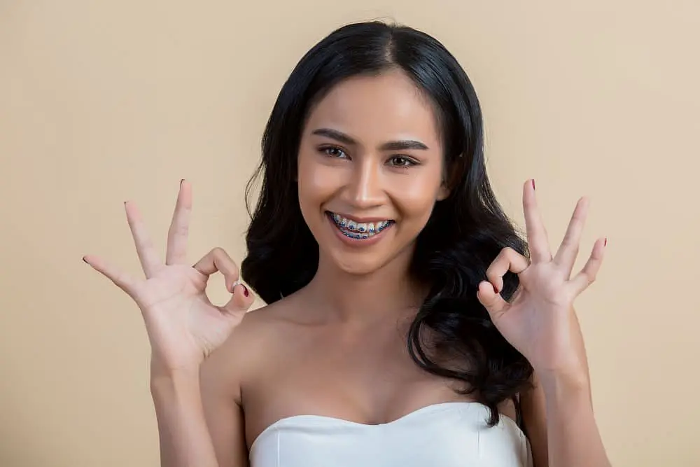 A woman with long black hair and traditional metal braces smiles and flashes an "OK" gesture with both hands in a photo for Holt Orthodontics in Twinsburg, OH.