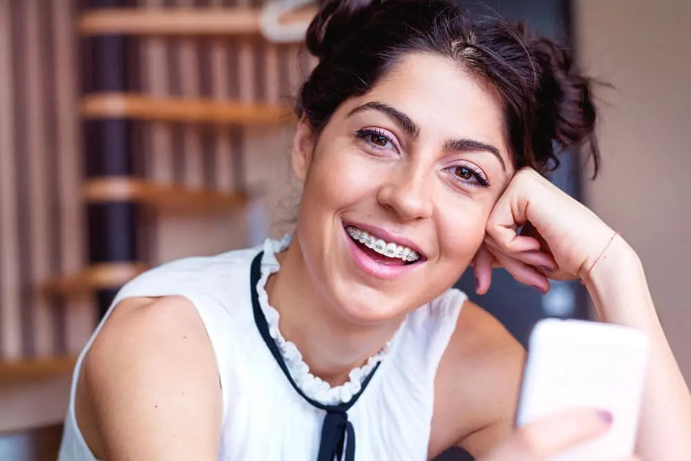 A young woman with dark hair in buns, wearing metal braces and a white blouse, smiles at the camera while holding her smartphone indoors at Holt Orthodontics in Twinsburg, OH.