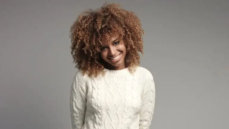 A person with curly hair and a white cable-knit sweater smiles subtly against a plain gray background at Holt Orthodontics in Twinsburg, OH.