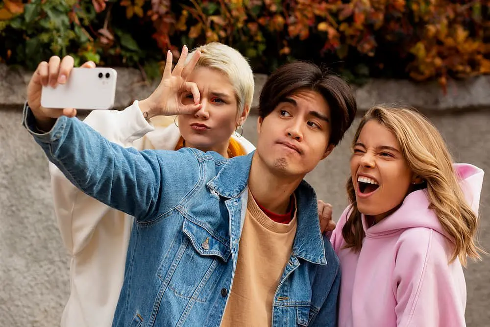 Three young adults pose for a selfie outdoors at Holt Orthodontics in Twinsburg, OH—one gives an "OK" sign, another proudly shows off their braces, and the third focuses on the phone.