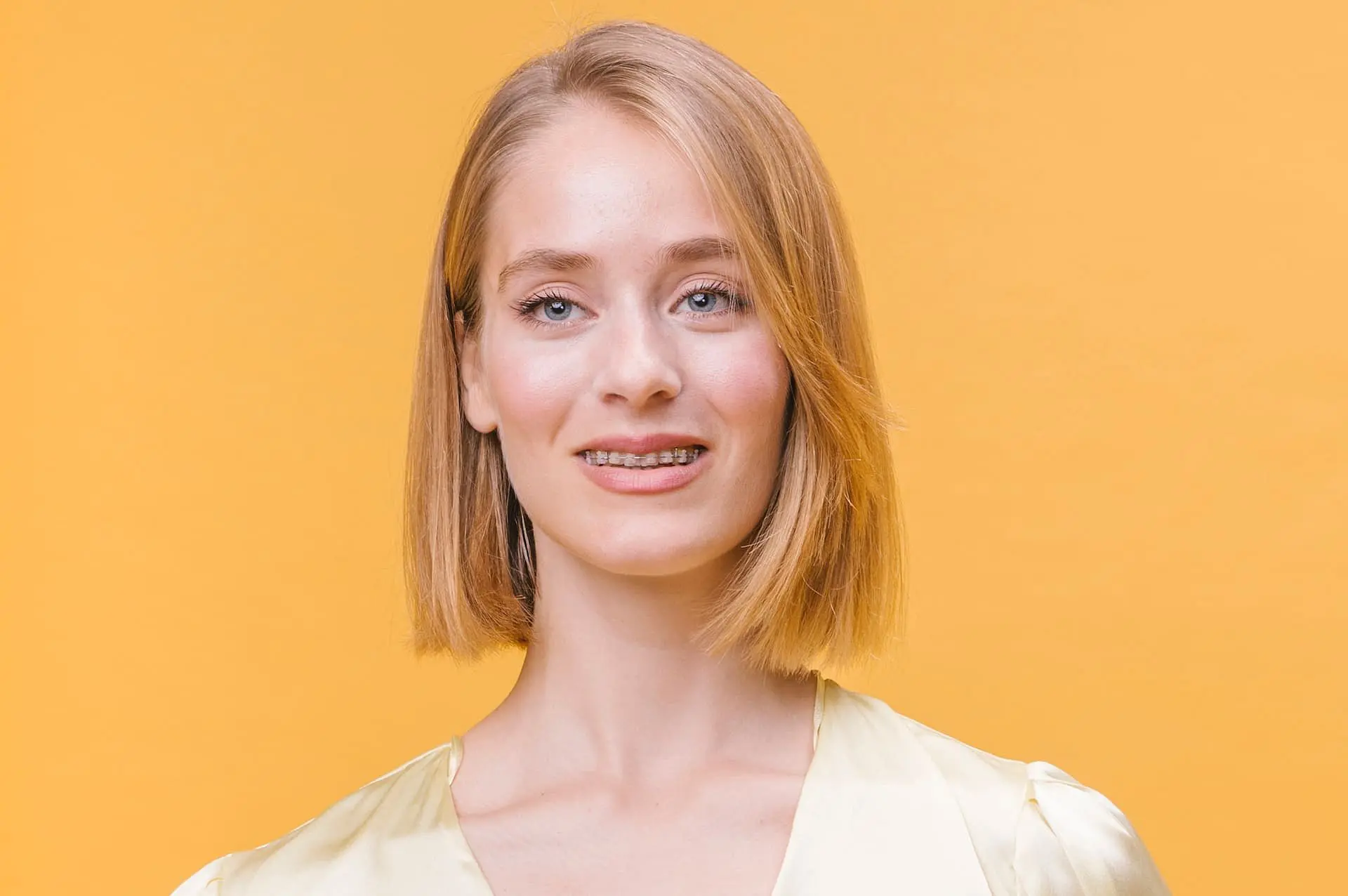 A woman with straight blonde hair and clear braces, dressed in a light yellow top, stands before a solid yellow background at Holt Orthodontics in Twinsburg, OH.