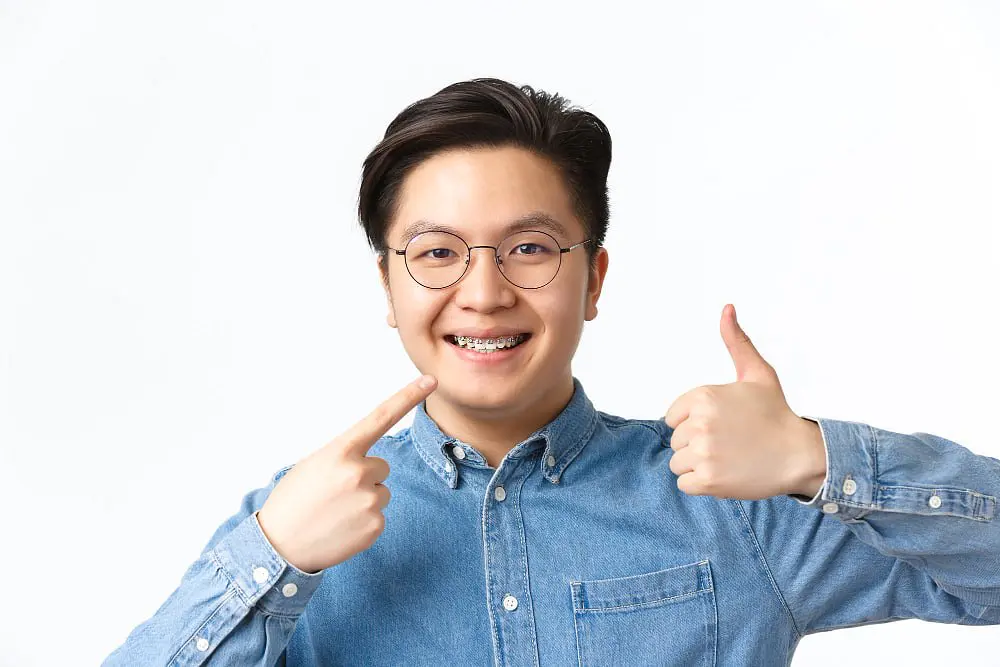 At Holt Orthodontics in Twinsburg, OH, a young person in glasses and a denim shirt smiles confidently, pointing to their dental braces and giving a thumbs up—highlighting the benefits of adult orthodontics.